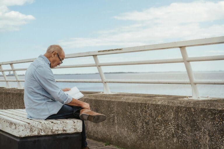 Side view of serious aged male wearing casual clothes and eyeglasses engaged in hobby reading book on embankment